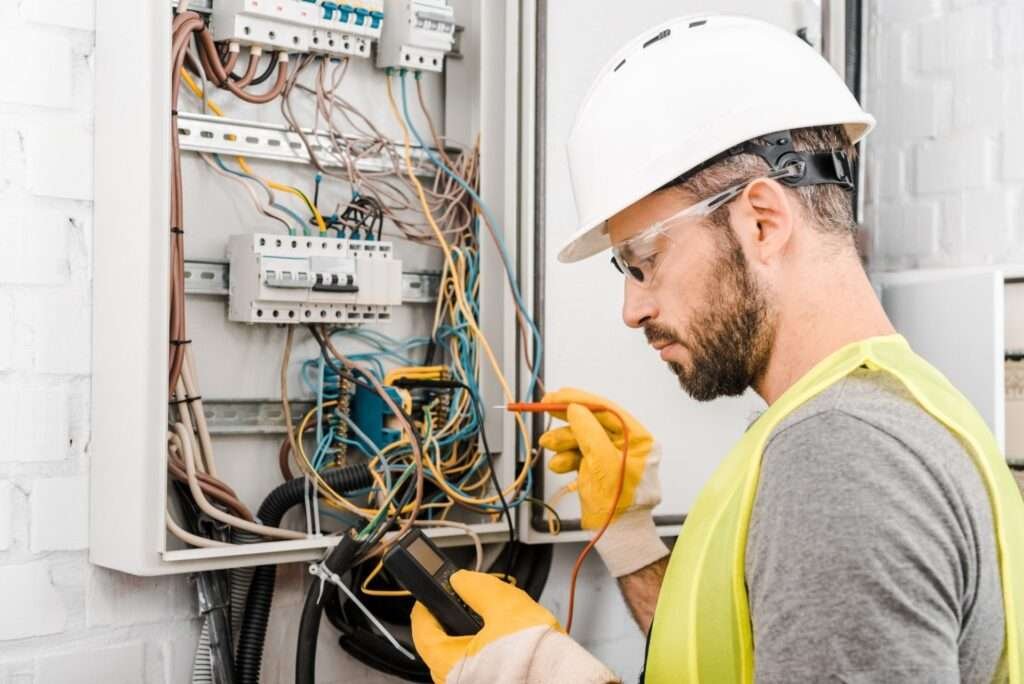 handsome electrician checking electrical box with multimetr in corridor e1624860078476